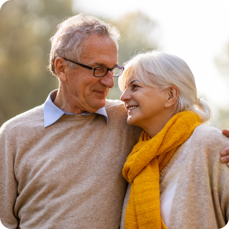 A couple in their 50’s outside, the man’s arm around the woman as they smile at each other.
